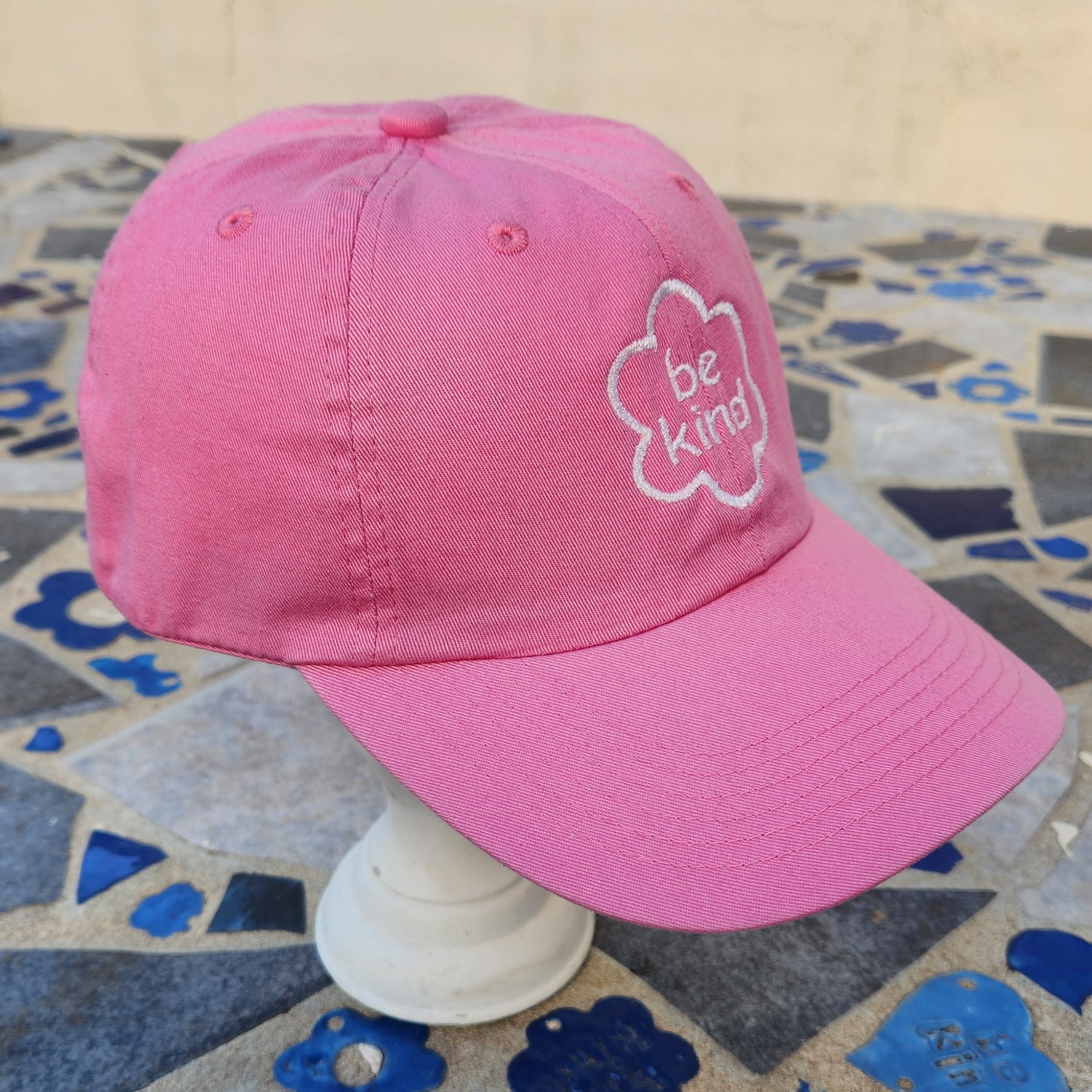 A Pink baseball cap with white embroidered flower on the front with text "be kind" in the middle